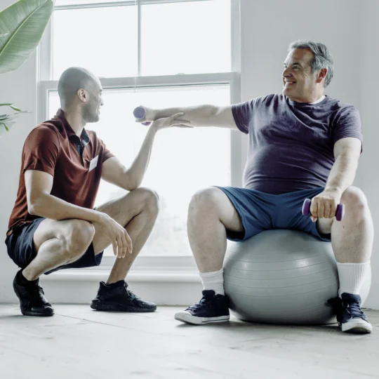 Älterer Herr auf Gymnastikball mit Trainer